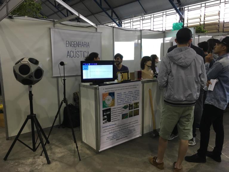 Foto do estande da engenharia acústica no descubra, tem algumas pessoas de pé olhando e dois estudantes atrás de um balcão.