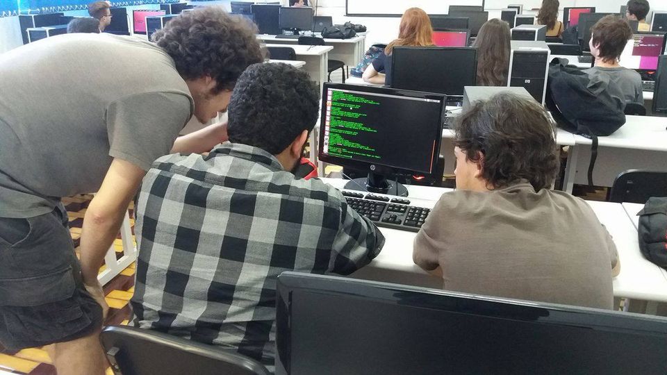 Três alunos brancos, olhando para tela de uma computador preta com letras verdes, perante outros alunos da aula