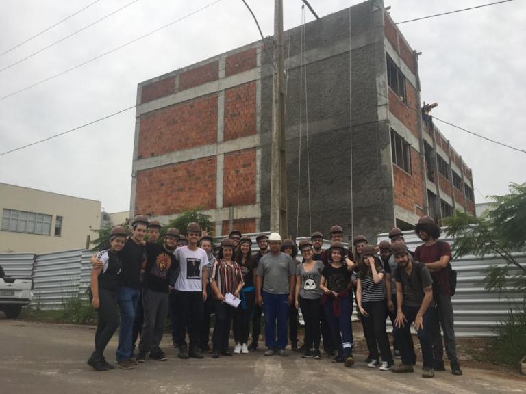 Foto de um grupo de pessoas com capacetes de obra posando para a foto e ao fundo um prédio em construção.
