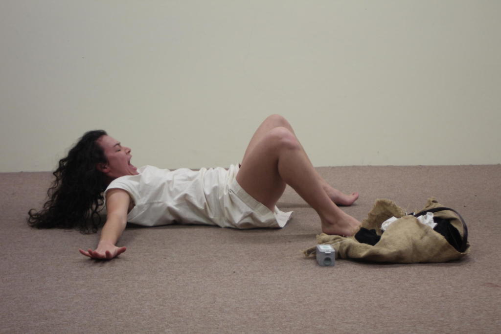 Mulher branca de cabelos cacheados, encenando uma peça deitada no chão de carpete bege. Em seu rosto, contém expressões de desespero.