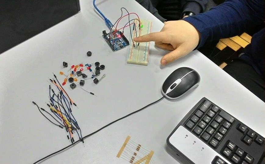 uma mão branca, junto de um teclado e mouse de computador, mexendo em alguns fios e peças variadas