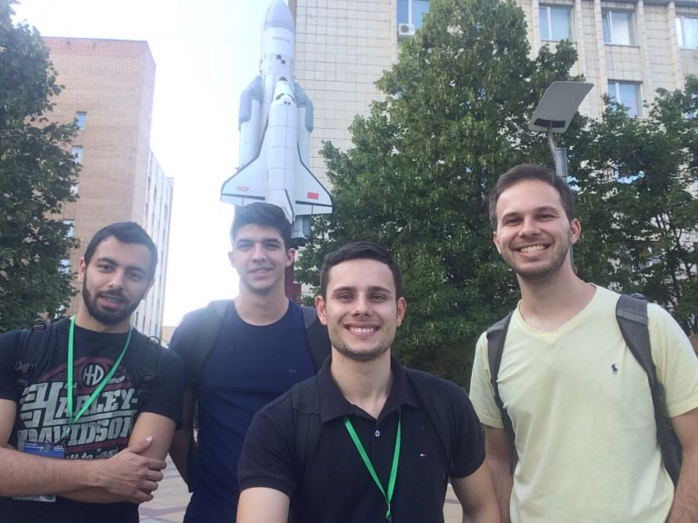 Quatro alunos do curso em um evento, na Rússia. Eles estão na rua e atrás tem um foguete