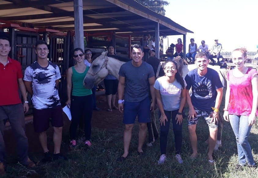Foto de alguns alunos em uma estrebaria com alguns cavalos.