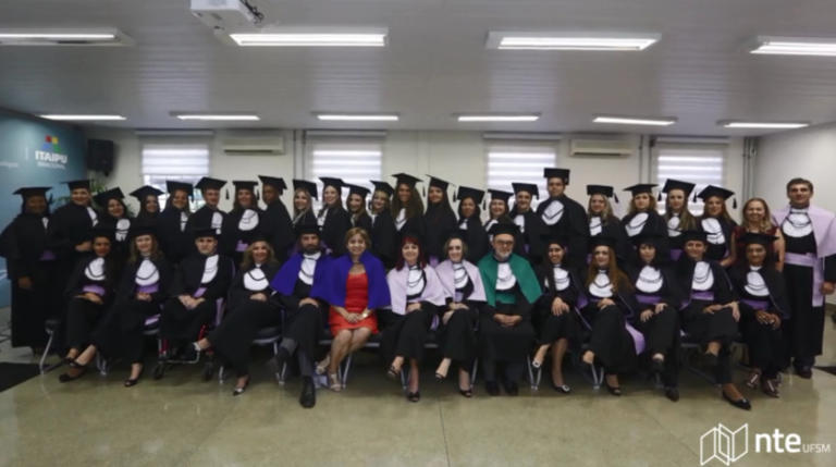 Alunos e professores com toga de formatura. Na primeirafileira as pessoas estão sentadas e na segunda estão de pé