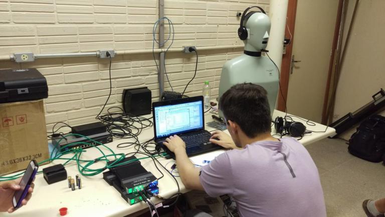 Foto de um menino de costas mexendo em um notebook que está sobre uma mesa cheia de fios e aparelhos e a metade de um manequim com fones de ouvido na cabeça.