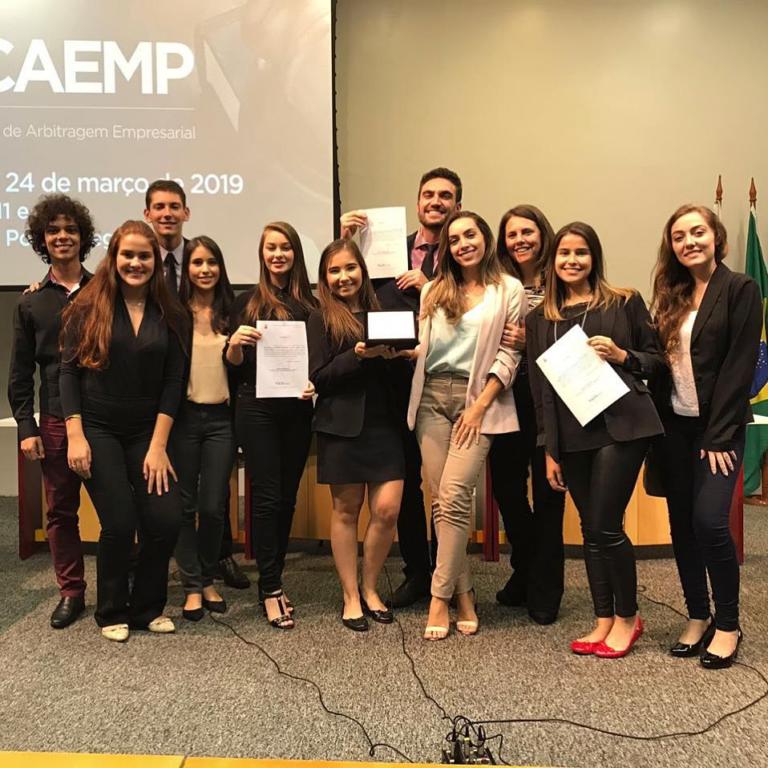 Foto de várias pessoas com roupas sociais, posicionadas lado a lados em frente de um telão.