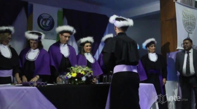 Uma bancada com professores e aluno vestindo toga de formatura. O aluno está de costas cumprimentando à todos