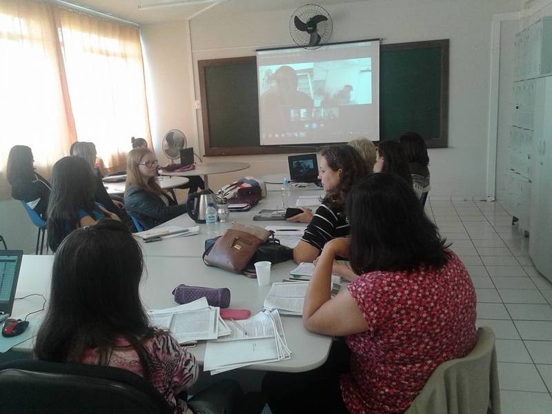 Foto de uma sala de aula com uma mesa redonda, e ao redor da mesa algumas mulheres que olham para um telão em sua frente.