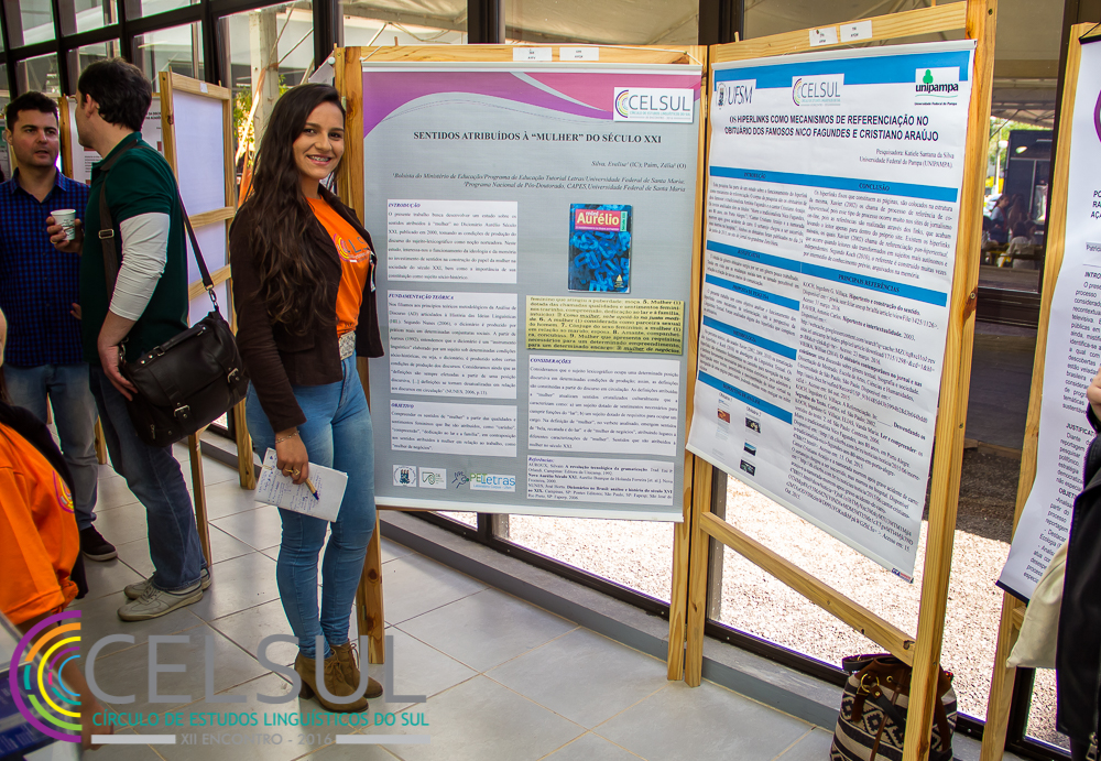 Foto de uma mulher posicionada ao lado esquerdo da foto, ela posa ao lado de um banner.