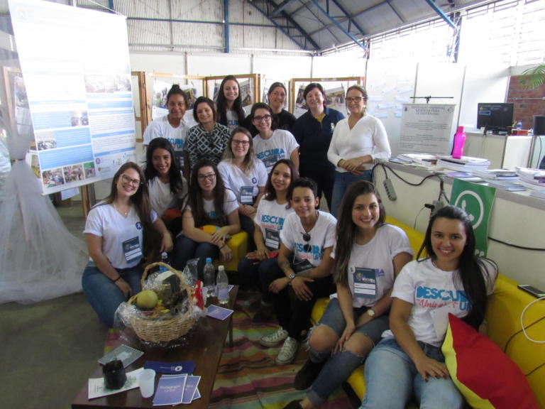 várias pessoas sentadas em um sofá amarelo e outras de pé atrás estão apresentando o estande do Descubra