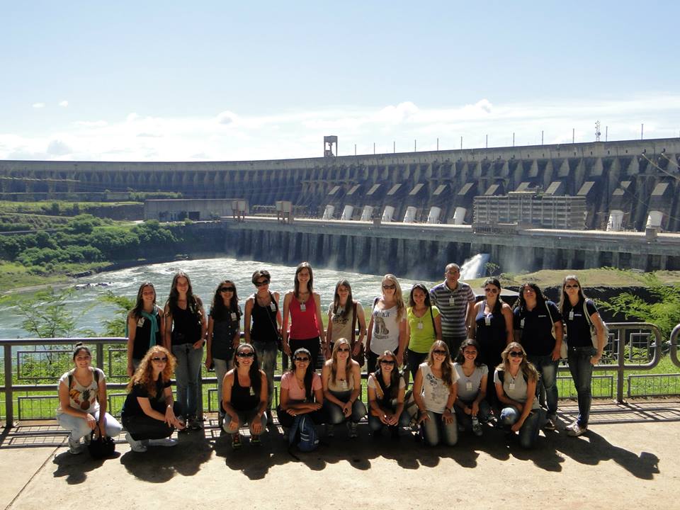 Foto de alunos posicionados lado a lado, estão sobre uma ponte, atrás deles tem a Usina Hidrelétrica de Itaipu.