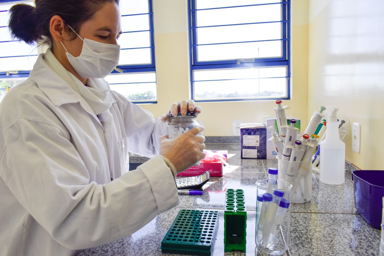 Foto de mulher usando máscara e jaleco brancos trabalhando em bancada de laboratório com diversos tubos de ensaio