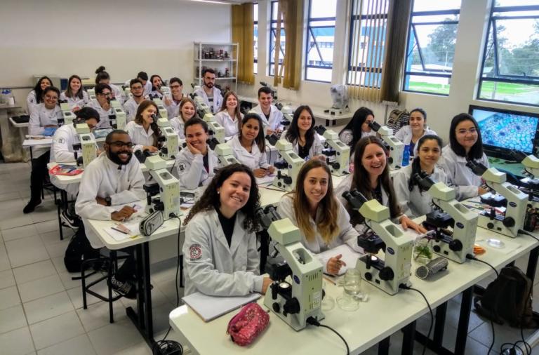 Foto de vários alunos de jaleco branco, sentados em fileira de mesas contendo um microscópio.