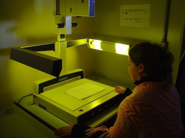 Mulher mexendo em uma máquina grande, analisando um documento. A luz da foto está amarelada