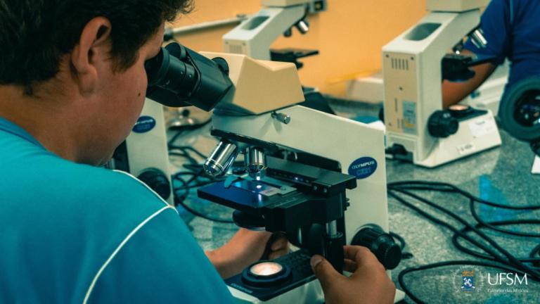 Homem de camiseta azul sentado olhando sobre um microscópio.