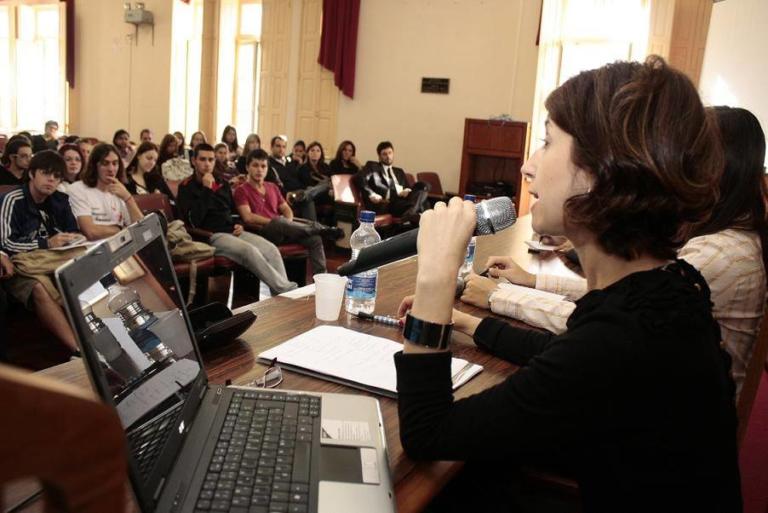 Foto de uma mulher sentada atrás de uma mesa falando em um microfone para várias pessoas.