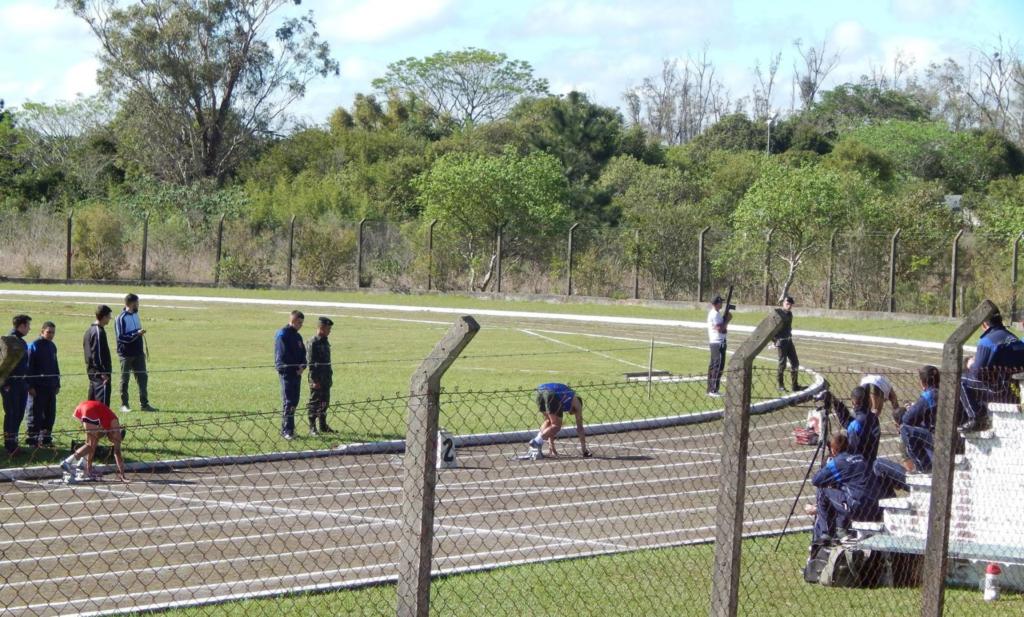 Foto da pista de atletismo, tem um homem se preparando para iniciar uma corrida e outras pessoas ao redor observando.