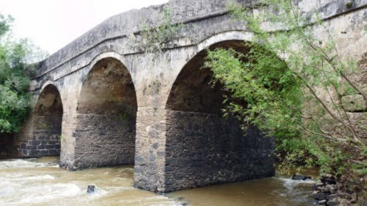 Ponte de pedra cinza com grandes arcos por onde corre o rio
