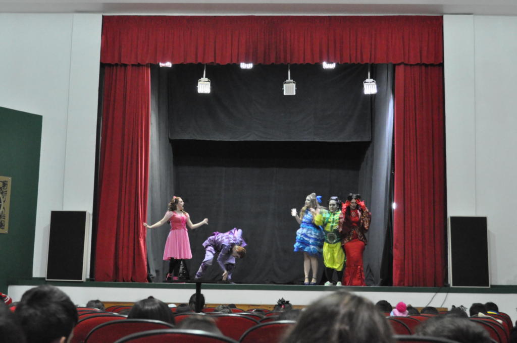 quatro mulheres e um homem branco, encenando uma peça, para uma plateia em um palco, que contém cortinas vermelhas e chão preto.