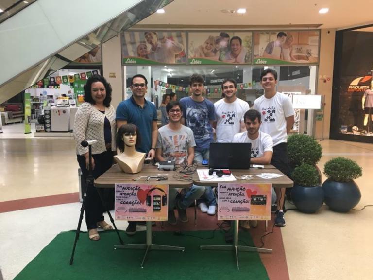 Foto de um grupo de pessoas, cinco estão de pé e dois sentados atrás de uma mesa que contem um notebook e a metade de um manequim.