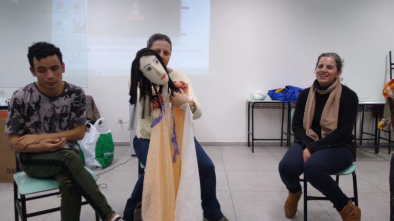 duas pessoas sentadas e uma em pé no centro com uma boneca.