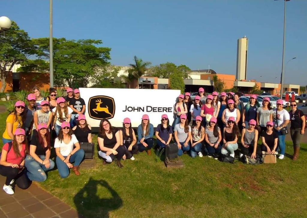 Foto de várias mulheres reunidas, posicionadas lado a lado em frente de um banner que tem escrito "John Deere", cada uma delas usa um boné rosa.