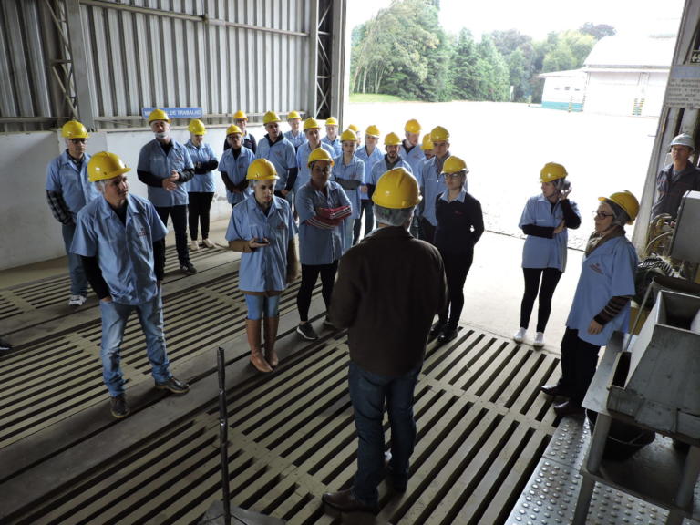 Foto de várias pessoas com jaleco branco e capacete amarelo. elas prestam atenção na fala de um homem que está a frente delas.