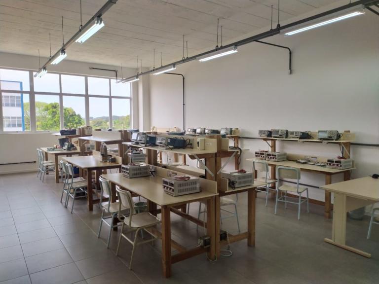 Foto de uma sala com mesas uma virada pra outra com cadeiras e equipamentos, é um laboratório.