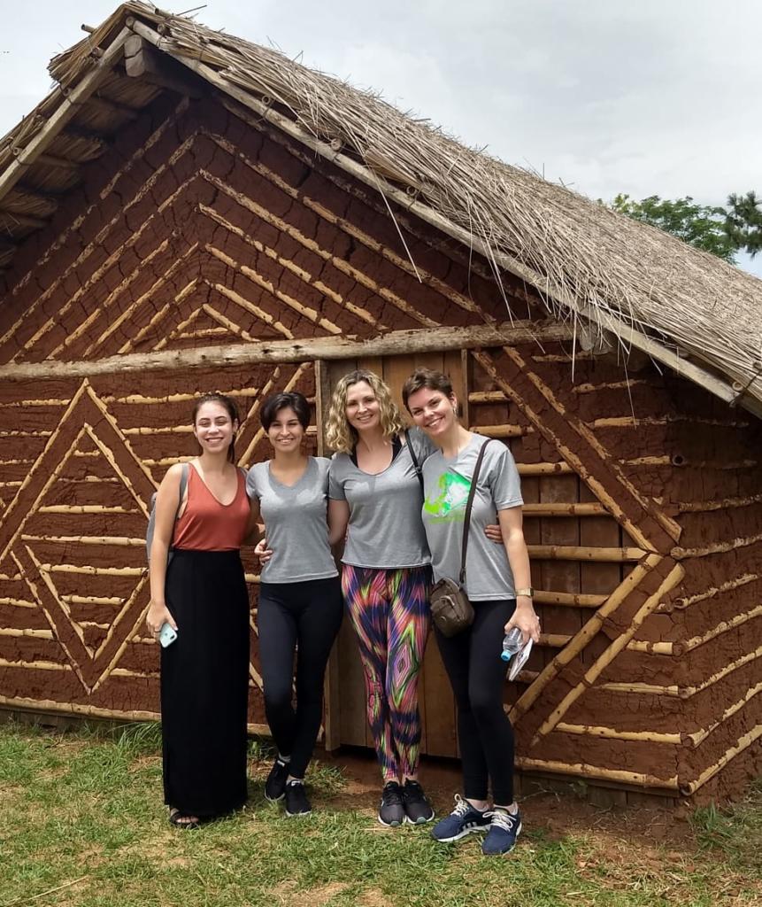 Quatro mulheres em frente a uma residência indígena feita de barro