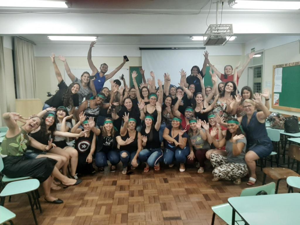 Foto de vários alunos em um sala reunidos para a foto, estão com as mãos levantadas, todos tem uma faixa verde na cabeça.