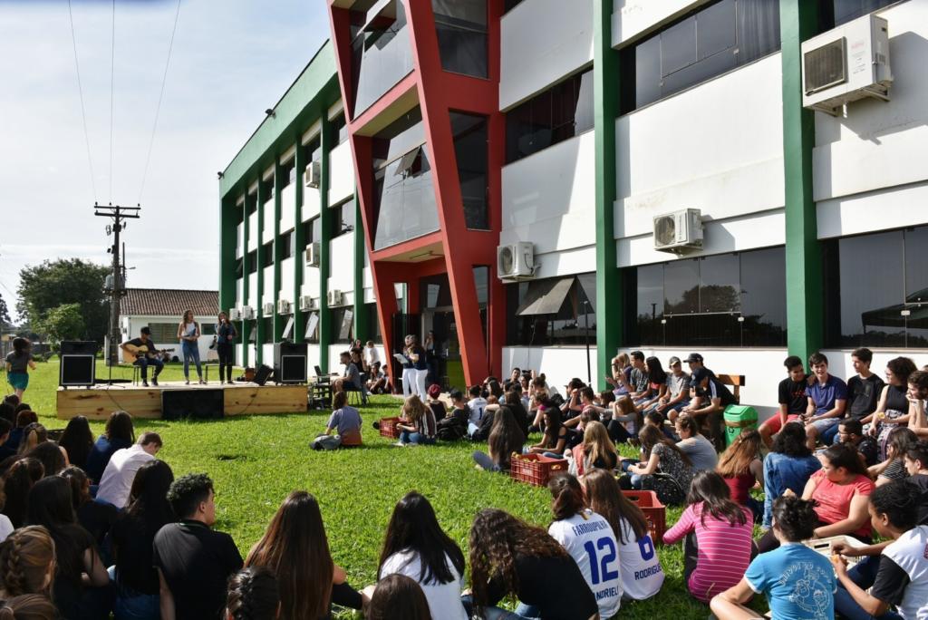 Pessoas sentadas no gramado em frente a prédio bege com verde e vermelho