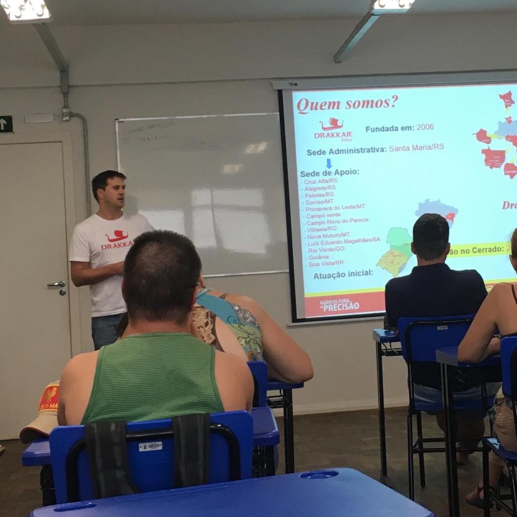 Foto de uma sala de aula, aparecem alguns alunos e o professor ao lado de uma apresentação de slides.
