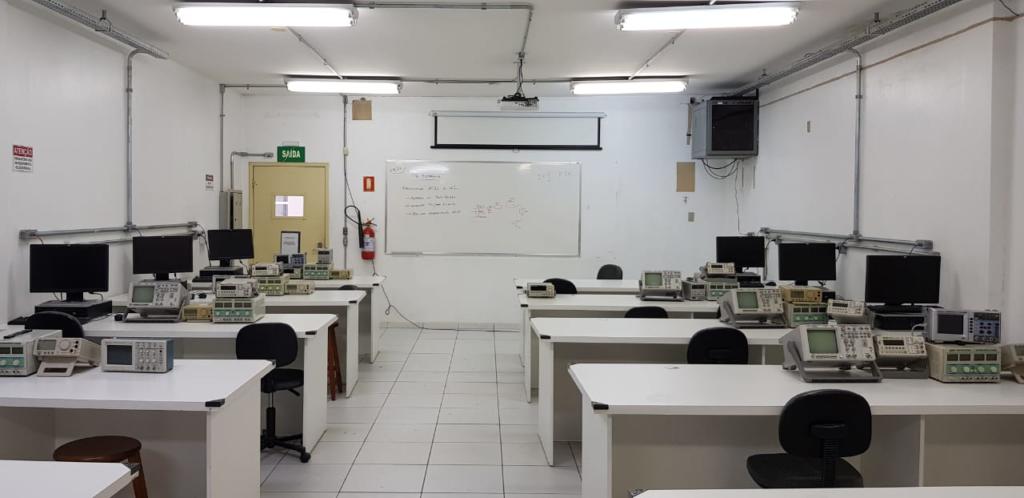 Na foto mostra um laboratório de paredes brancas com varias mesas retangulares com computadores em cima