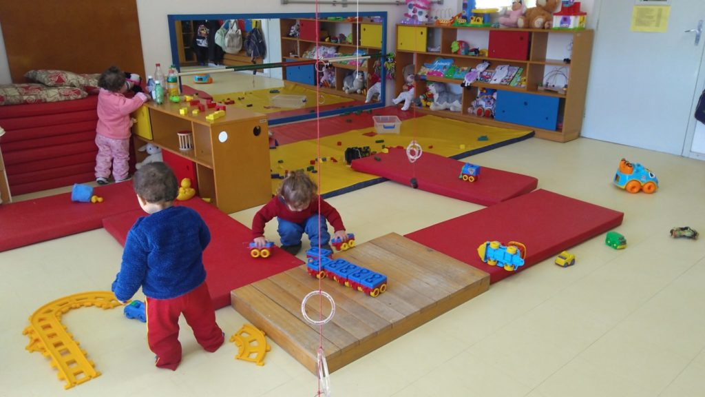 Crianças brincando em sala com diversos brinquedos, tapetes
