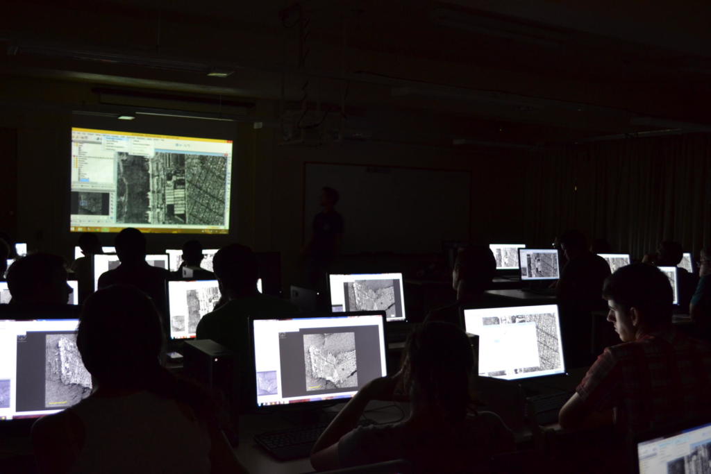 Foto de uma sala de aula com a lu apagada, nela tem alunos sentados em seus computadores olhando imagens de radar.