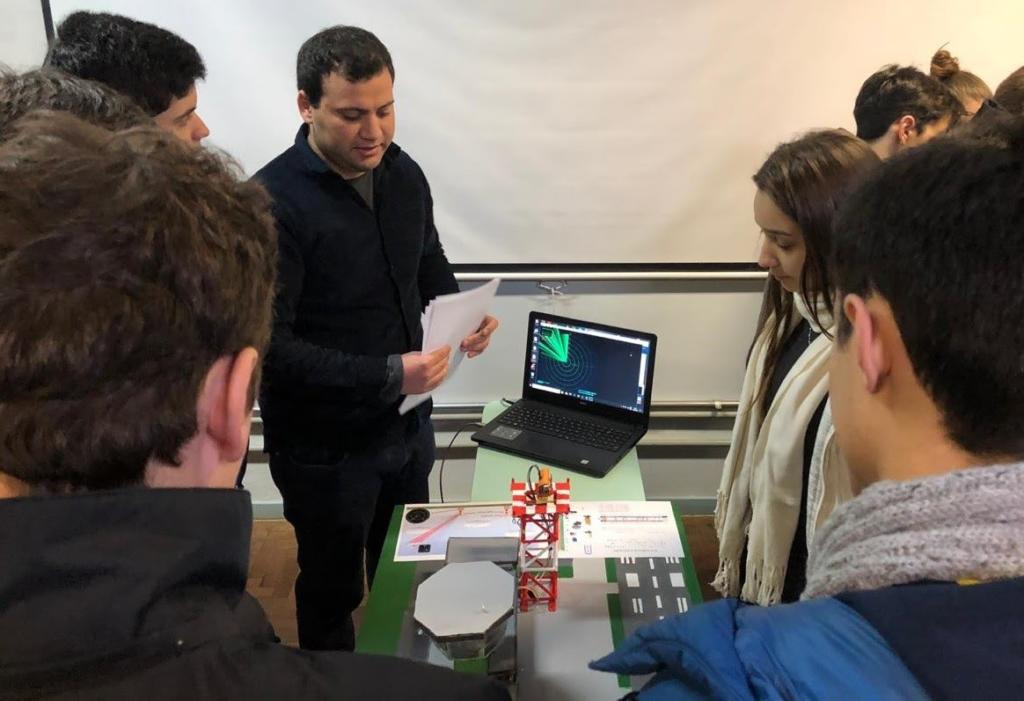 Diversas pessoas em torno de bancada com equipamentos, uma pessoa apresentando a experiência para os demais