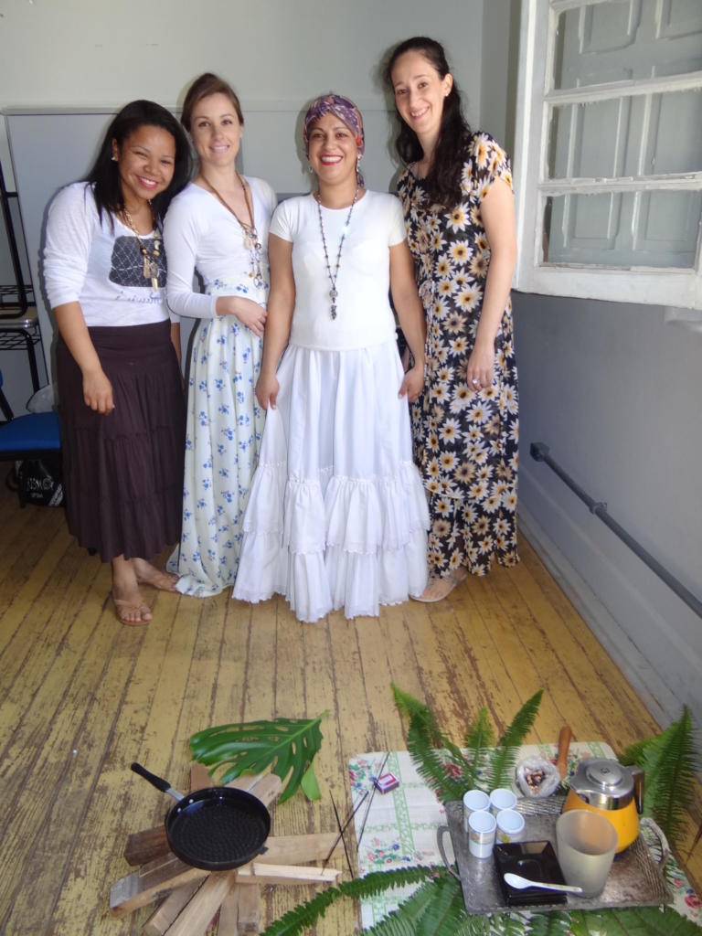 4 mulheres em pé, uma delas veste traje baiano branco. Na frente uma arrumação de plantas e chá no chão