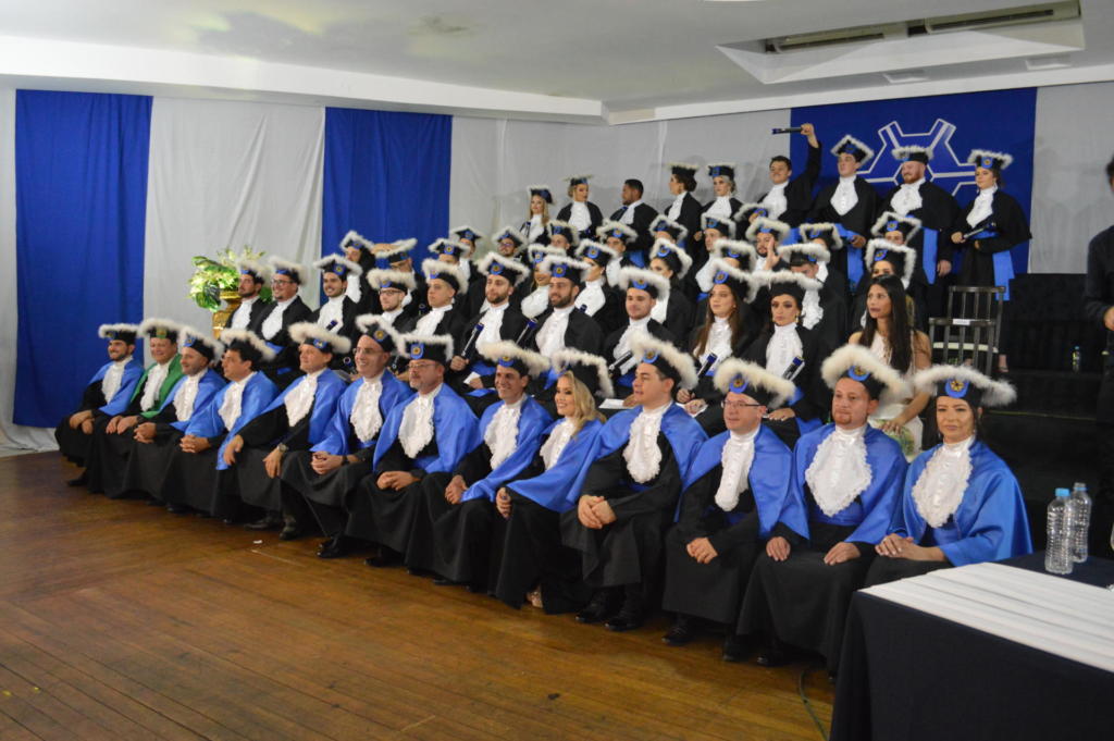 Alunos e professores vestindo becas de formatura e capelo sentados num palco