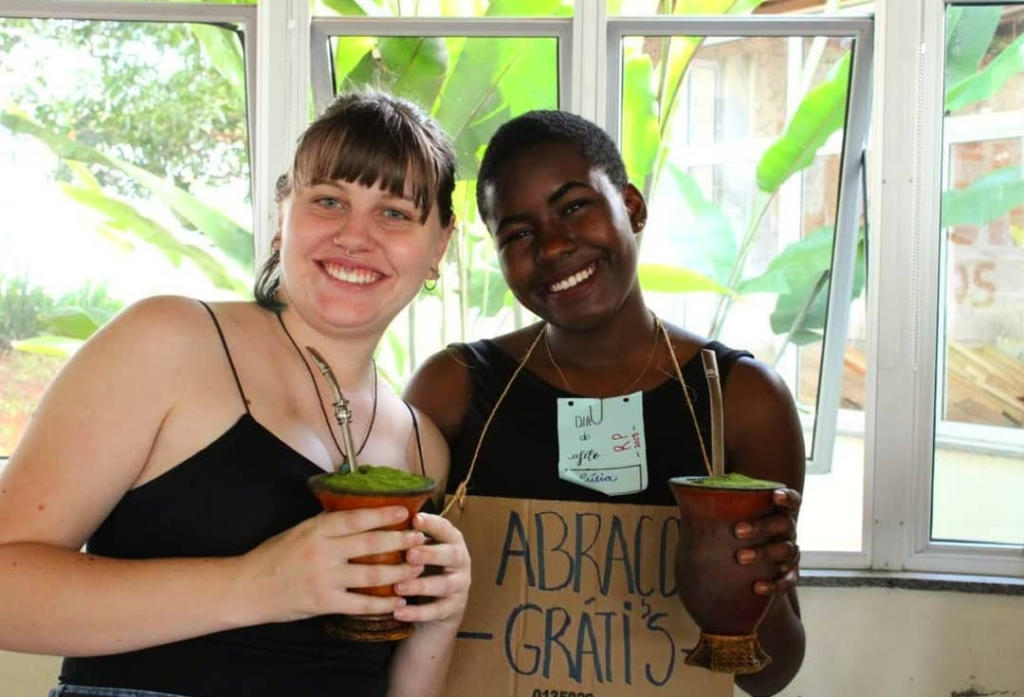 Duas mulheres abraçadas tomando chimarrão, uma tem um cartaz no peito com a frase Abrações Grátis