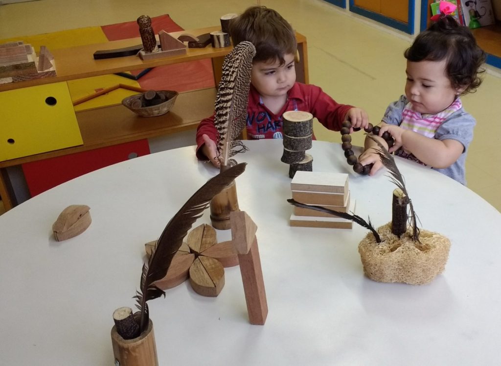 Duas crianças sentadas em mesa mexendo em brinquedos de madeira