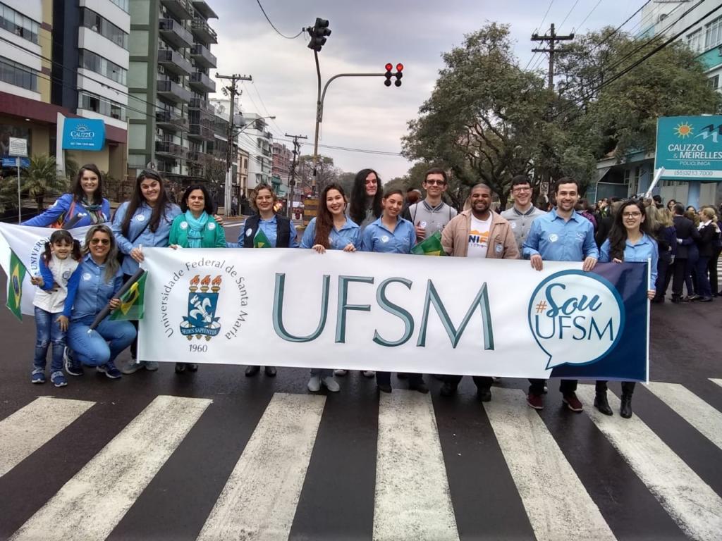 Várias pessoas na frente de uma faixa de pedestres segurando uma faixa que diz UFSM e Soiu UFSM.