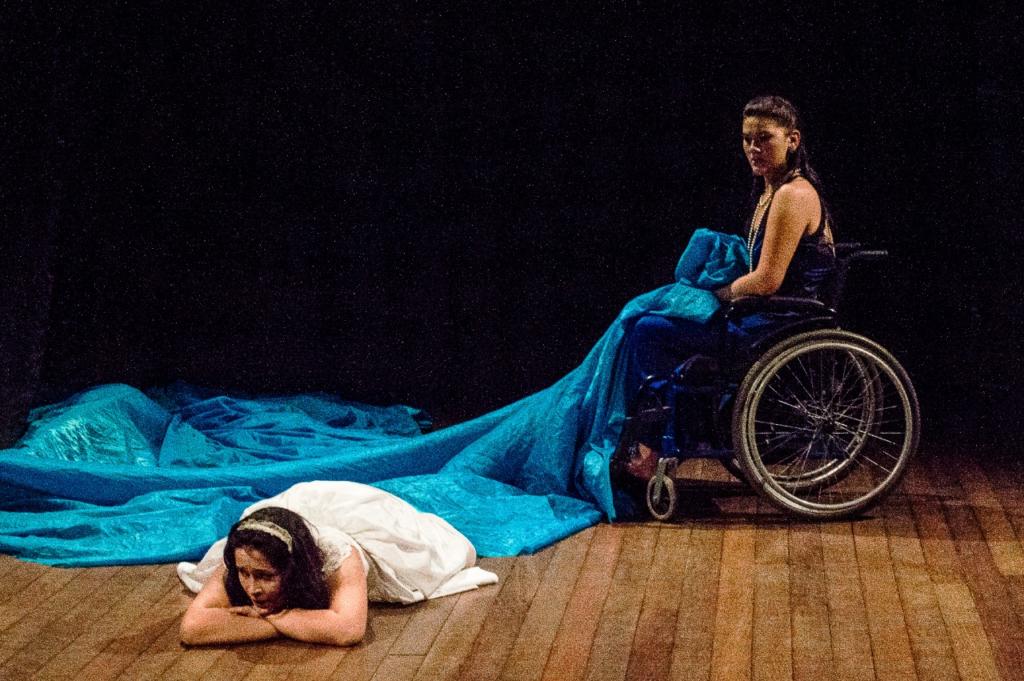 mulher de branco deitada no chão, dela sai um tecido azul carregado por uma mulher em cadeira de rodas