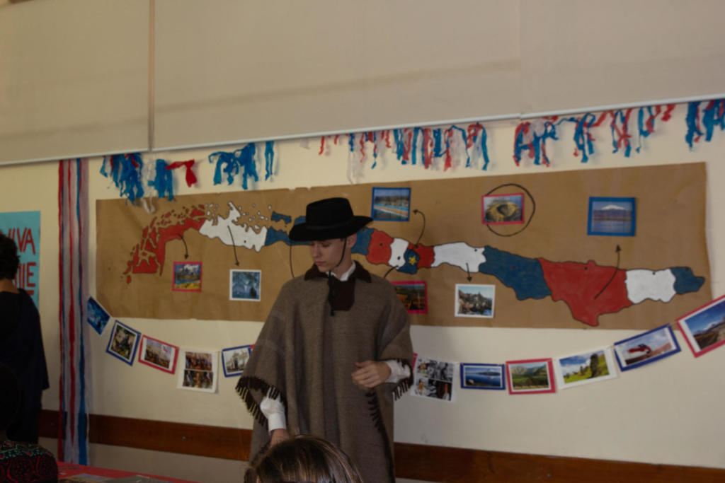 Homem vestido de pala marrom e chapéu preto falando na frente da sala de aula