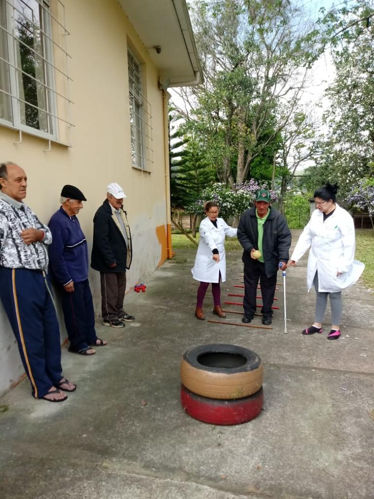 Foto de quatro idosos e duas cuidadoras, elas incentivam um dos idosos a se aproximar de dois pneus empilhados.