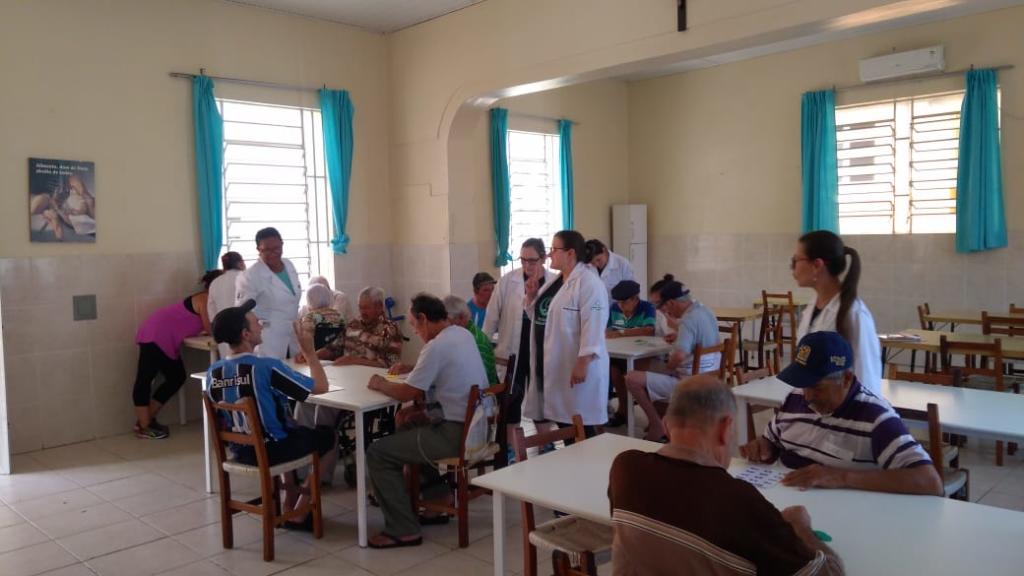 Foto de uma sala bem aberta, com algumas mesas e cadeiras, alguns idosos estão sentados nessas cadeiras enquanto jogam um bingo que as cuidadoras fizeram.