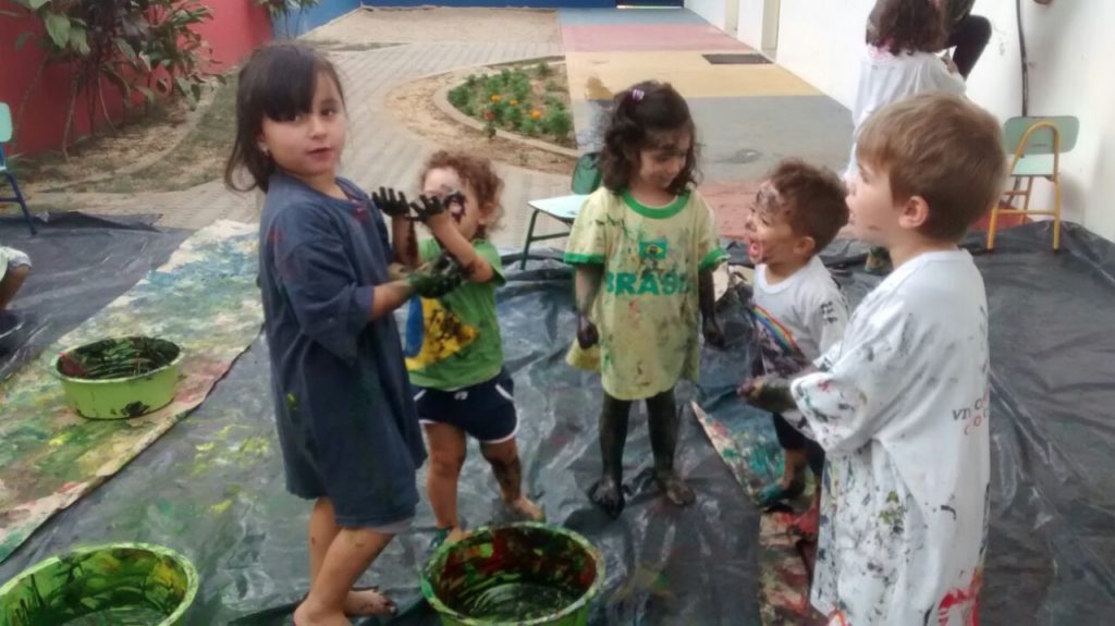 Na foto há cinco crianças sobre uma lona preta. Todas as crianças estão brincando com tinta guacha. As tintas estão misturadas dentro de bacias rasas de cor verde