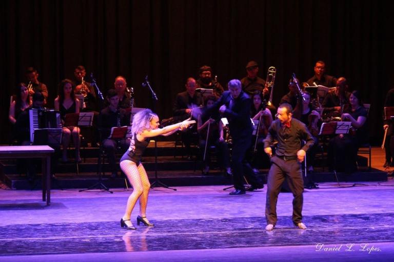 Homem e mulher dançando em palco, orquestra tocando ao fundo