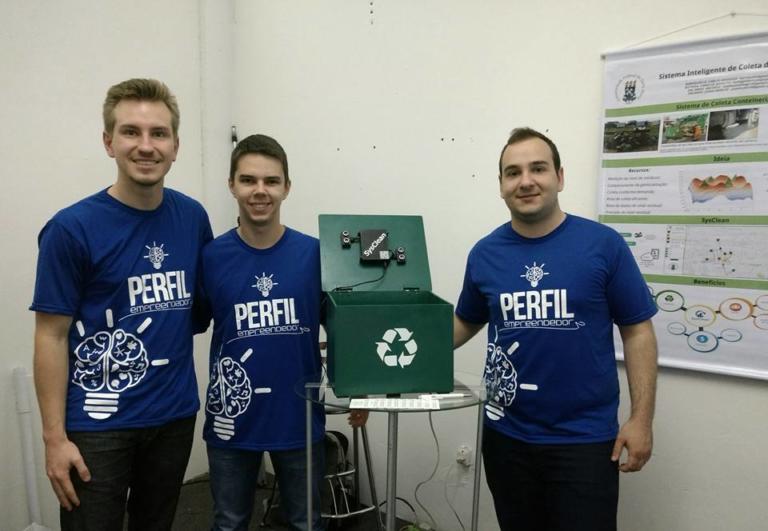 Três alunosvestindo azul e no centro uma caixa com o simbolo de reciclagem verde