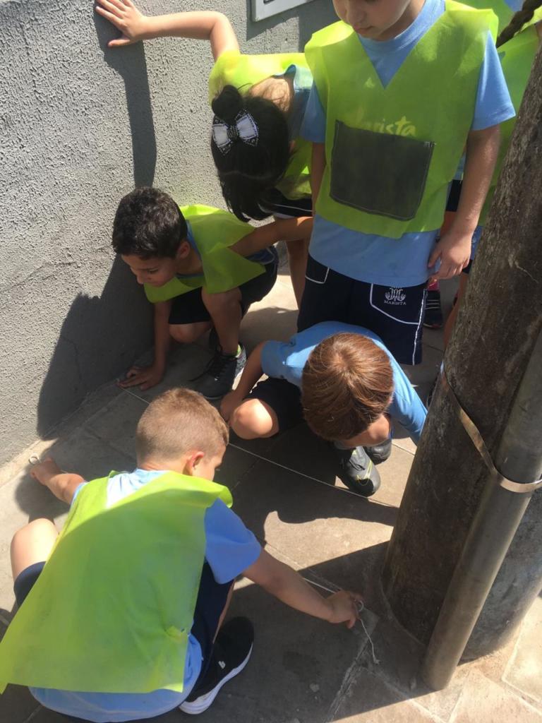 Na foto possui crianças brincando acocadas em um chão de cimento. Todas usam bermudas pretas, camiseta azul e colete verde