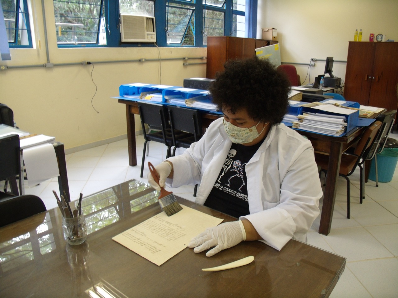 Pessoa vestindo jaleco, máscara e luvas brancas manipula documento sobre mesa